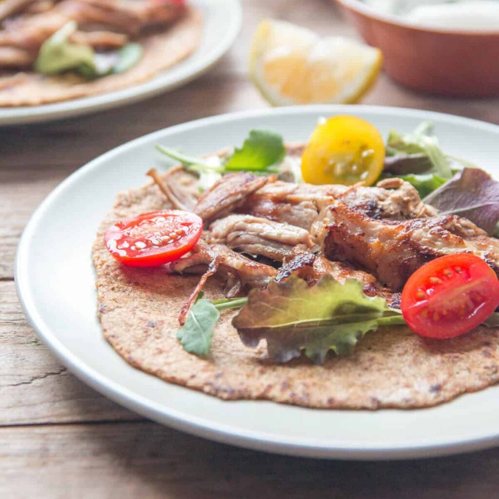 Closeup of plates with crispy pork carnitas with sliced open cherry tomatoes and lettuce on a flaxseed tortilla.