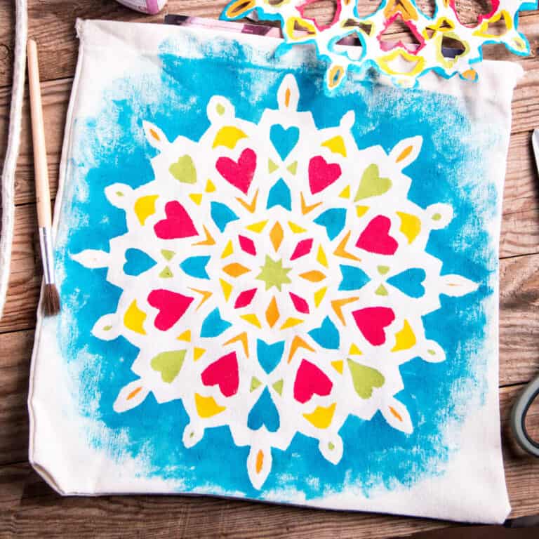 A fabric tote bag features a colorful mandala-like stencil design with red, yellow, and green shapes on a blue background, displayed on a wooden surface.