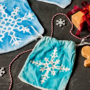 Blue drawstring pouches with white snowflake designs are displayed on a dark surface, along with snowflake ornaments and orange soap bars tied with red ribbon.